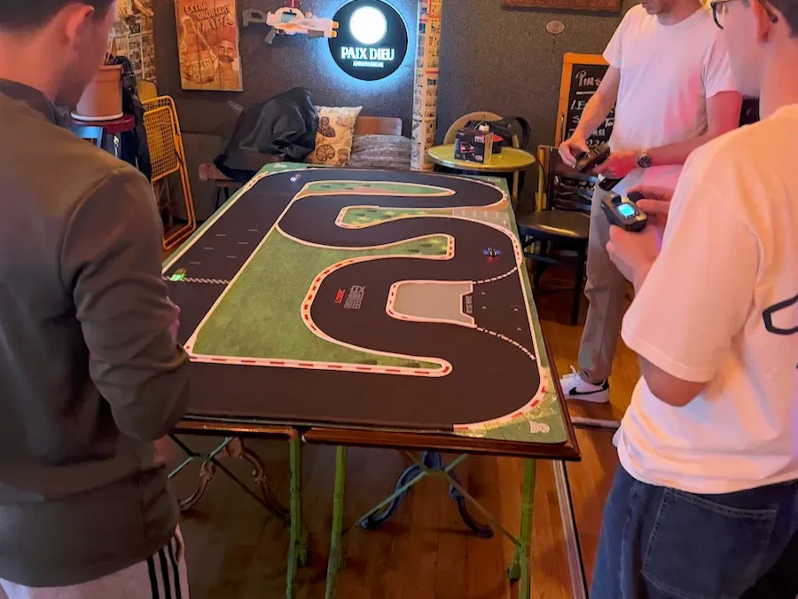 Quatre pilotes debout autour d’un circuit RC 1/76 posé sur une table, radiocommandes en main